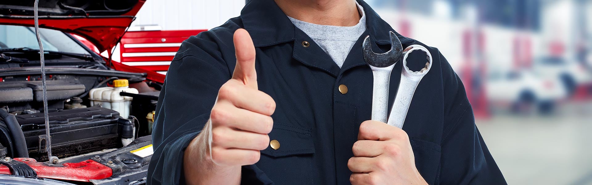 An auto mechanic holding wrenches and giving a thumbs-up gesture in front of an open car hood in a repair shop service bay.